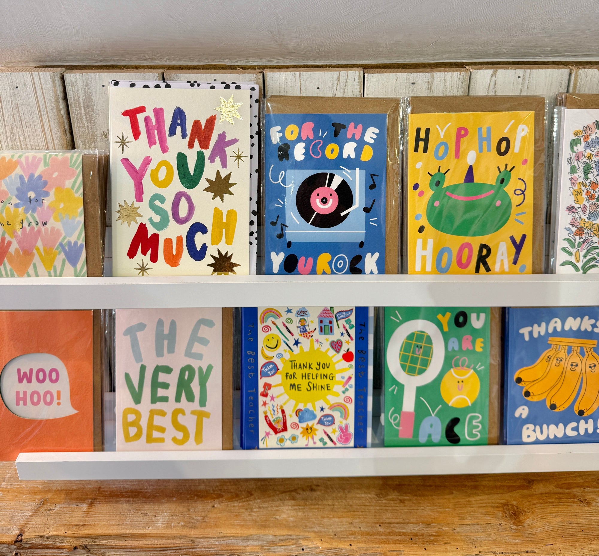 A row of colourful greeting cards displayed on white shelves. The cards feature playful, hand-drawn illustrations and humorous phrases such as 'THANK YOU SO MUCH', 'FOR THE RECORD YOU ROCK', 'HOPHOP HOORAY', 'THE VERY BEST', and 'WOO HOO!'.